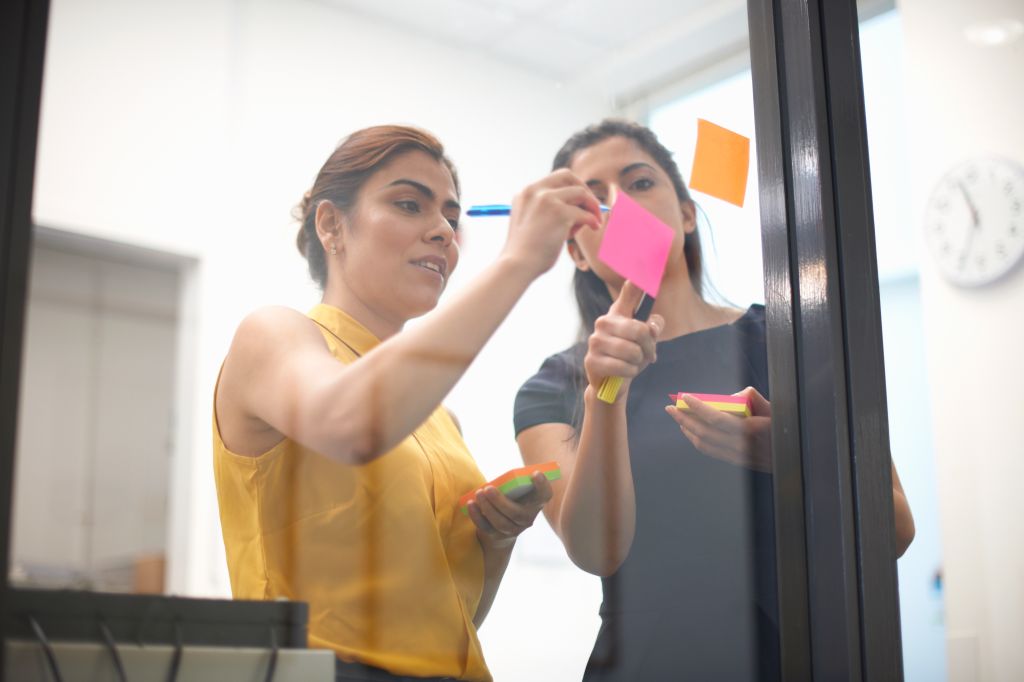 Two businesswomen writing out sticky notes on office glass wall