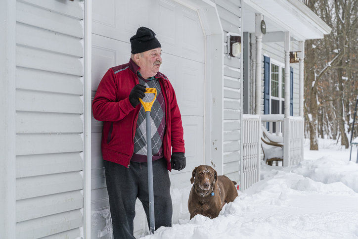 A tired senior man with a shovel takes a break when cleaning the snow in front of his garage. His dog makes a company.