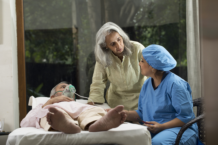 Female nurse explaining to woman while old male patient is lying on bed