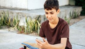 Teen boy sitting on skateboard using his smartphone