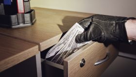 Man hand in a black glove takes money out of an office desk drawer in the night light of a flashlight