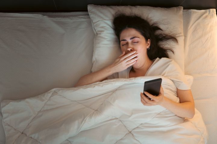 Top view of young Caucasian woman using smartphone and yawning, lying in bed at night. Insomnia, sleepless and social media addiction
