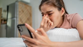 Exhausted Asian young woman hurt eye while using mobile phone on bed. Attractive girl lying down on bed, feeling visual fatigue and eye strain tired from overwork and massage dry irritable eye at home
