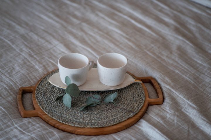 On the Linen bedspread lies a wooden round tray, on it are two empty ceramic cups, green eucalyptus