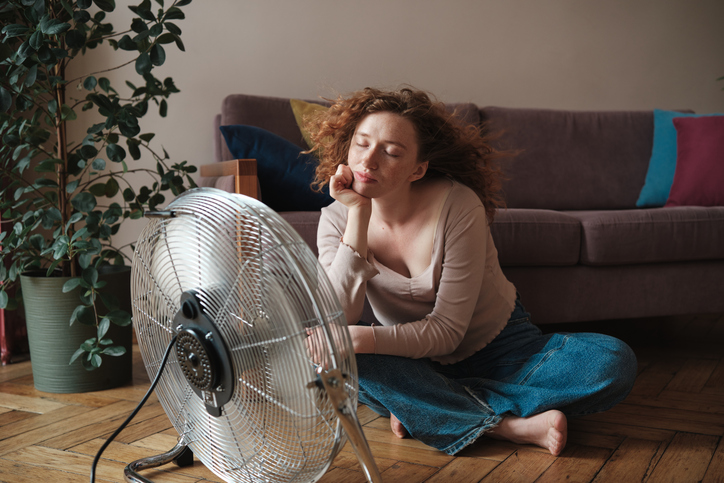 Woman suffering from summer heat at home using electric fan