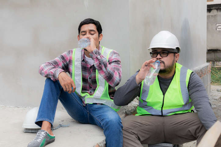 Two Asian builder workers wearing safety reflected vest and helmet tired thirsty hydrated drinking water from plastic bottle together relaxed sitting on construction site on overheated sunny day