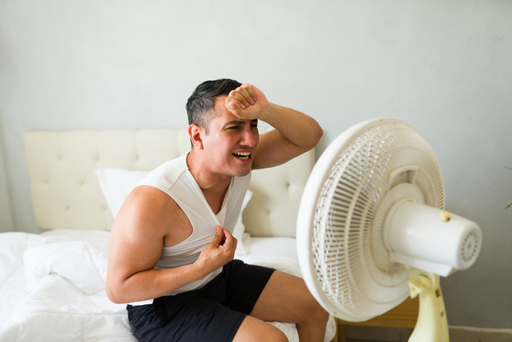 Sweaty man suffering from summer heat, feeling hot and exhausted in his bedroom while sitting on bed and using an electric fan