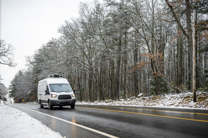 Commercial cargo mini van with refrigerator unit on the roof transporting cargo running on the wet slippery winter road with snowy forest on the side