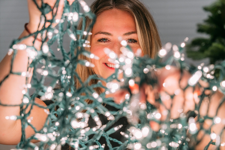 Smiling Portrait of a Cheerful Young Woman Holding Christmas Lights while Decorating a Christmas Tree