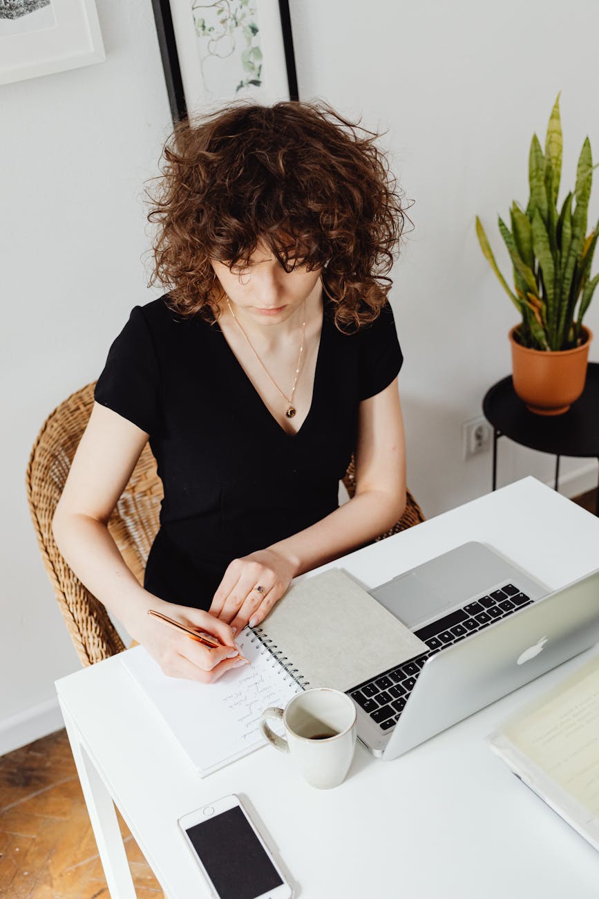 high angle shot of a woman writing on a notebook near a laptop