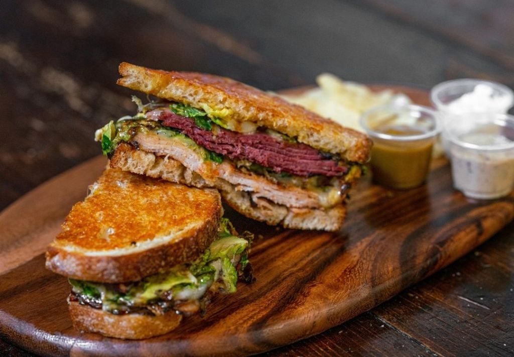 A close-up view of a grilled sandwich on a wooden board, featuring layers of meat, vegetables, and melted cheese between slices of toasted bread.