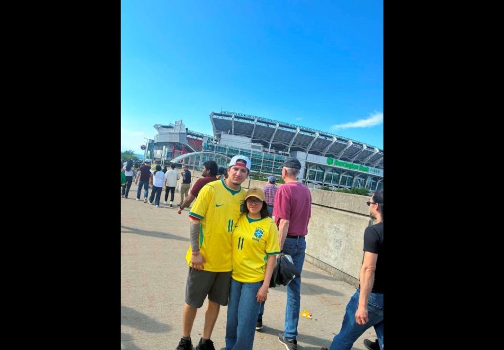 A group of people standing in front of a large sports stadium with a clear blue sky overhead. The stadium appears to be the home of the Brazilian national soccer team, as evidenced by the jerseys worn by the people in the image.