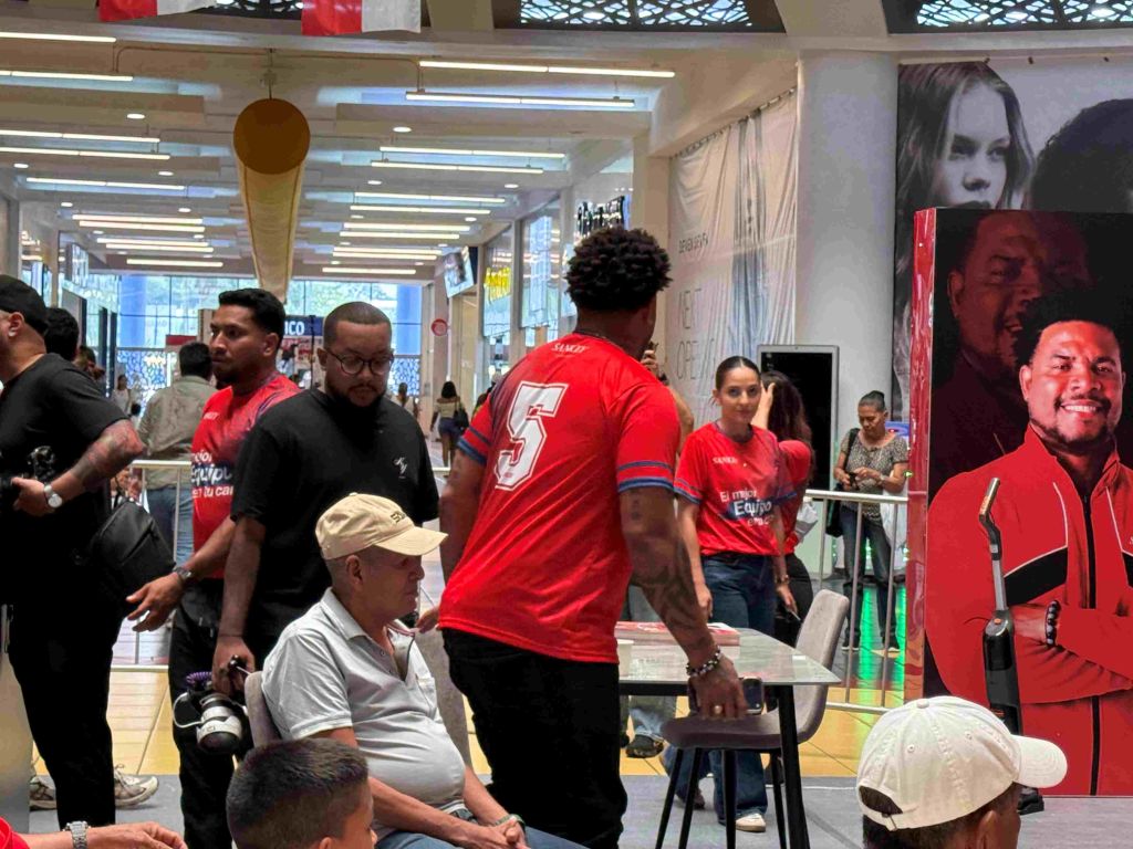 Crowded indoor space with people wearing red sports jerseys, some with jersey number 5, and others in casual clothing. Visible signs and advertisements in the background.