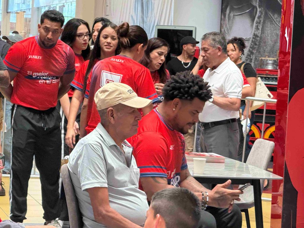 A group of people wearing red shirts with the text "El mejor en wi" gathered around a table, with an older man in a gray shirt and a younger man in a red shirt.