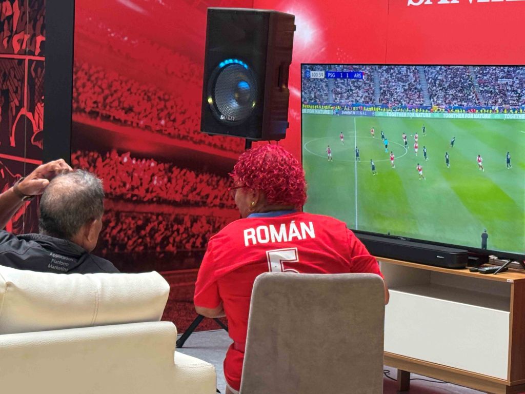 A person wearing a red jersey with the name "ROMAN" watching a soccer match on a large screen in a room with red walls and a crowd of people.