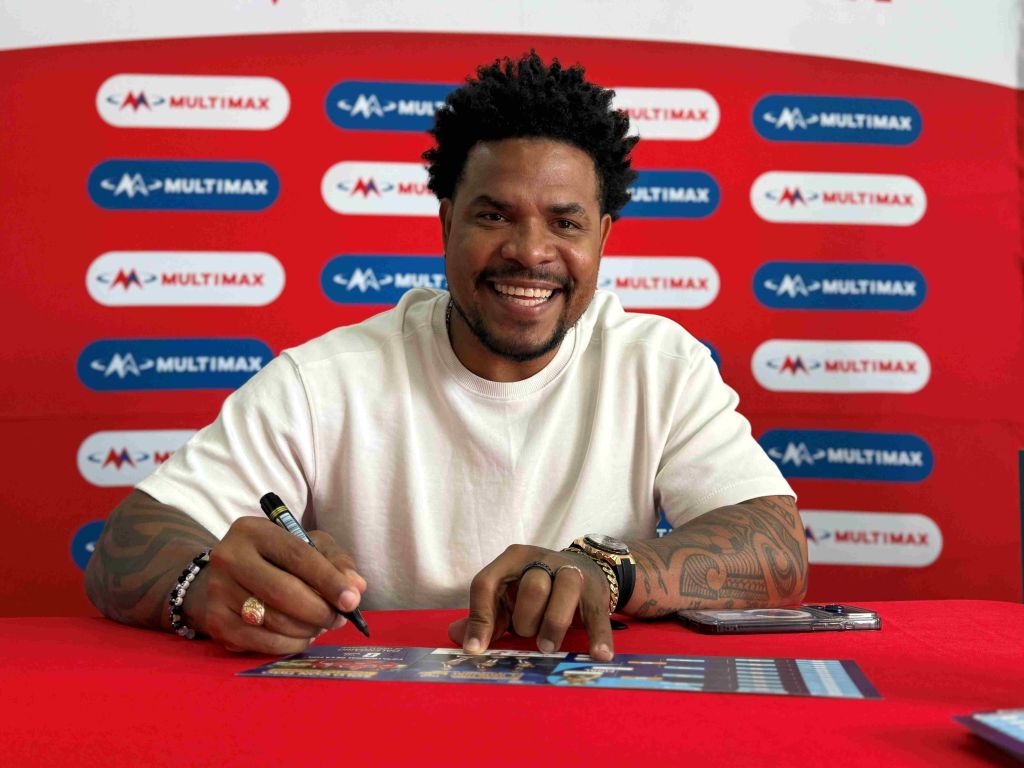 A smiling man with curly hair and tattoos sitting at a desk with Multimax branding in the background.