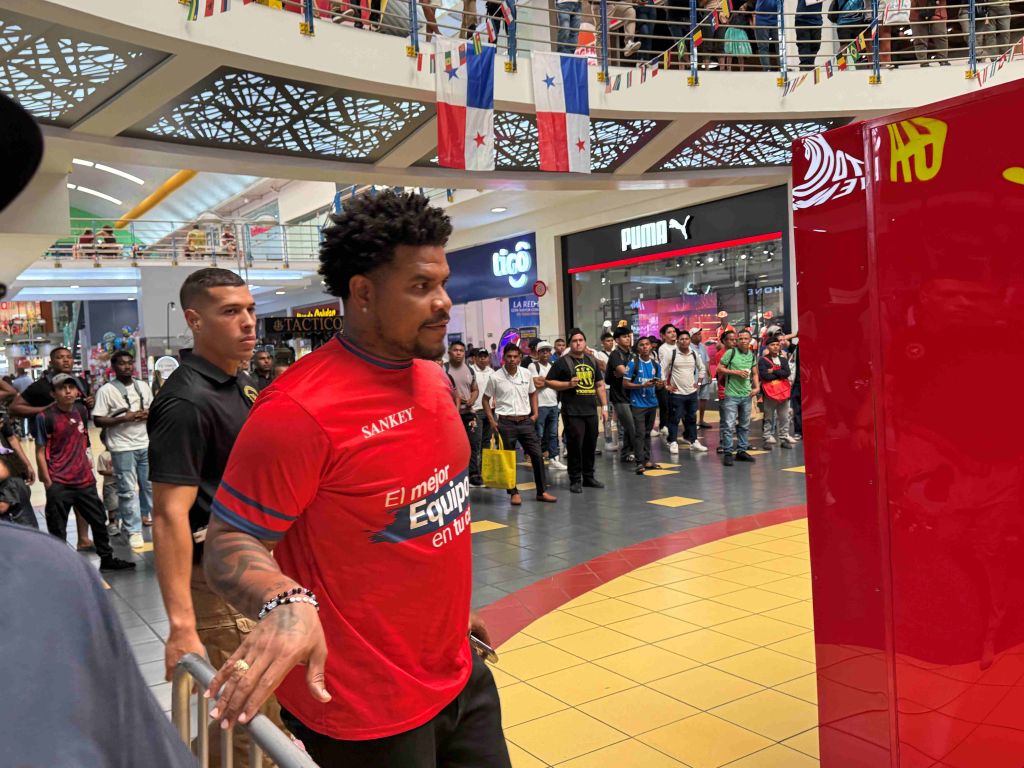 A man wearing a red jersey with the text "Sankey" and "El mejor equipo en tu" stands in a crowded indoor shopping mall with flags and banners hanging overhead.