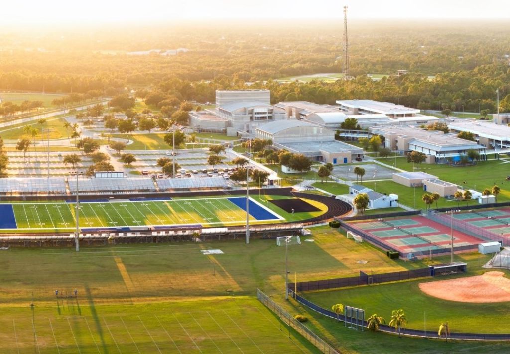 Aerial view of a college campus with sports facilities, including a football field, baseball diamond, and tennis courts, surrounded by trees and buildings under a golden sunset.