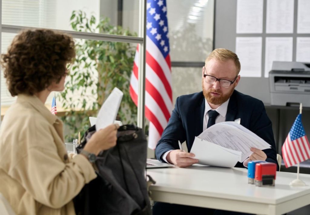 Two people in an office setting, one seated at a desk reviewing documents, the other standing and holding a folder. An American flag is visible in the background.