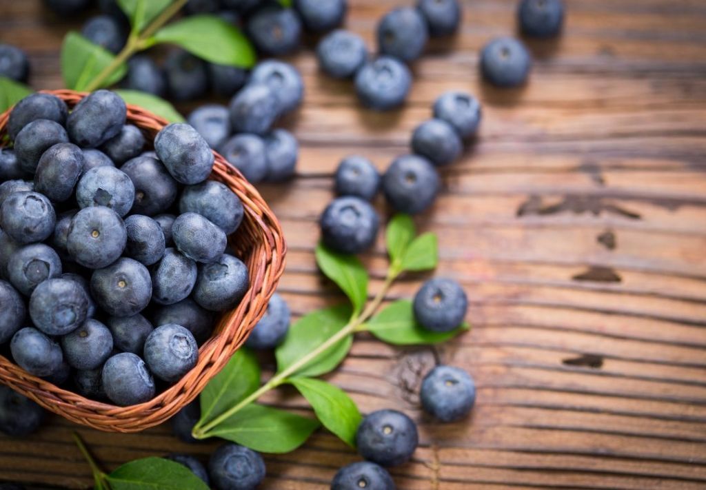 A wicker basket filled with fresh, ripe blueberries on a wooden table, surrounded by green leaves.