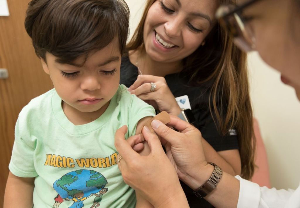 A young child wearing a "Magic World" t-shirt is being comforted by a smiling adult, likely a parent or caregiver, in a medical setting.