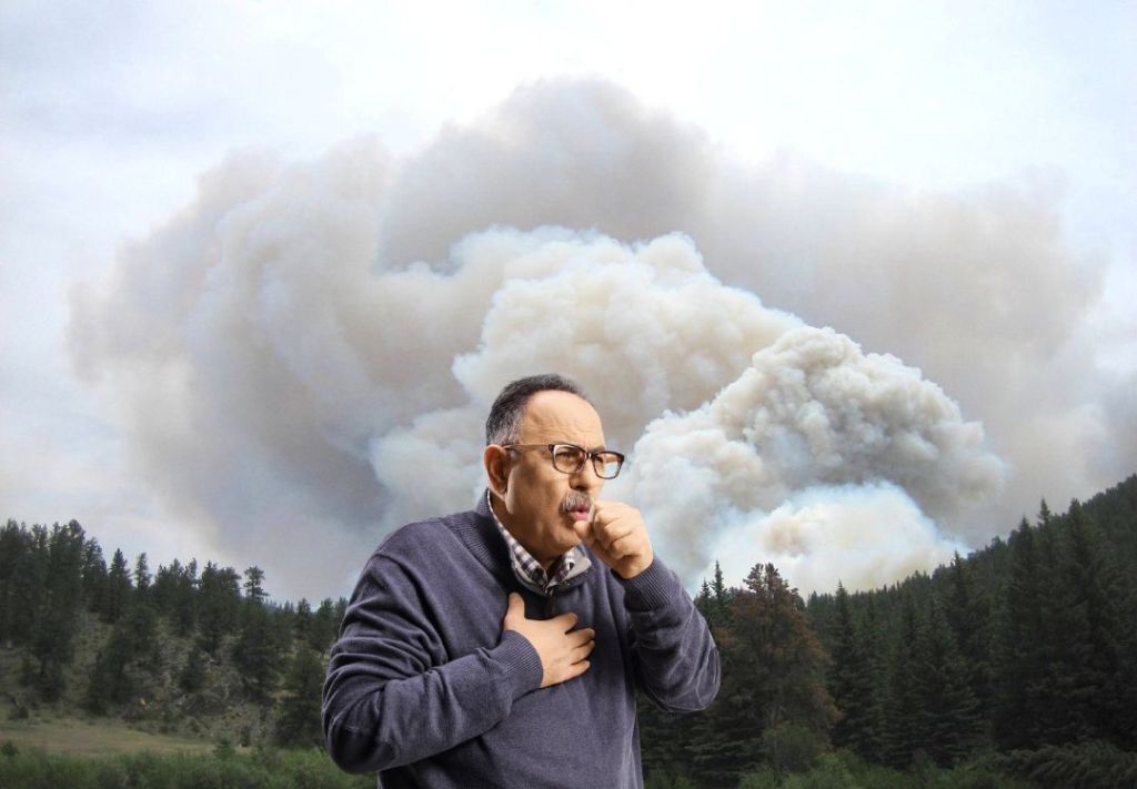 A man in glasses and a sweater stands in front of a large cloud of smoke from a wildfire in a forested area.