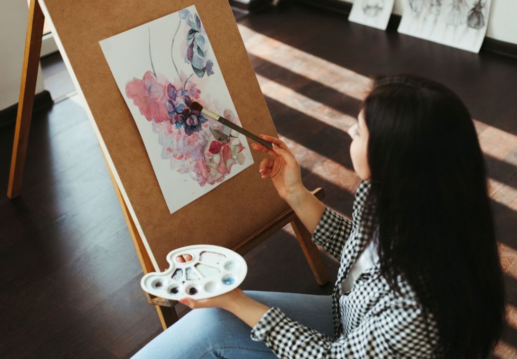 A person in a checkered shirt painting a watercolor floral artwork on an easel in a room with wooden furniture.