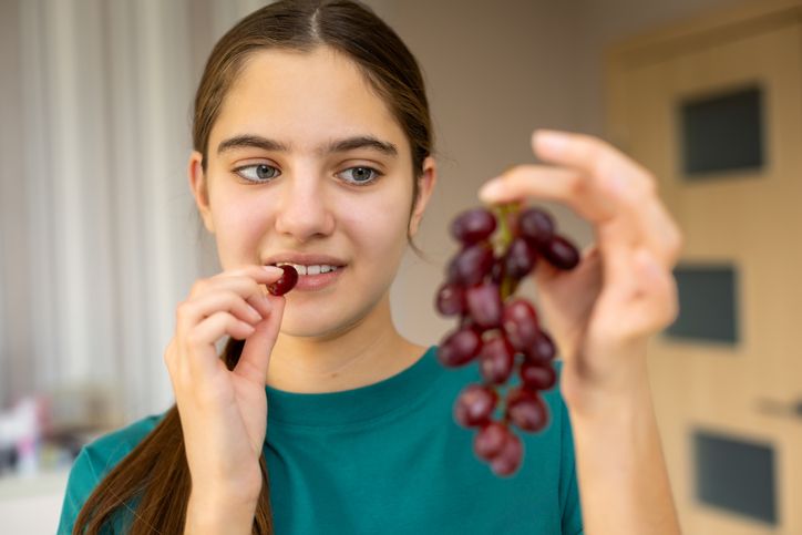Young Woman Eating Red Grapes Indoors. Young woman in a teal shirt holding a bunch of red grapes and bringing one grape to her mouth in a softly lit indoor setting.