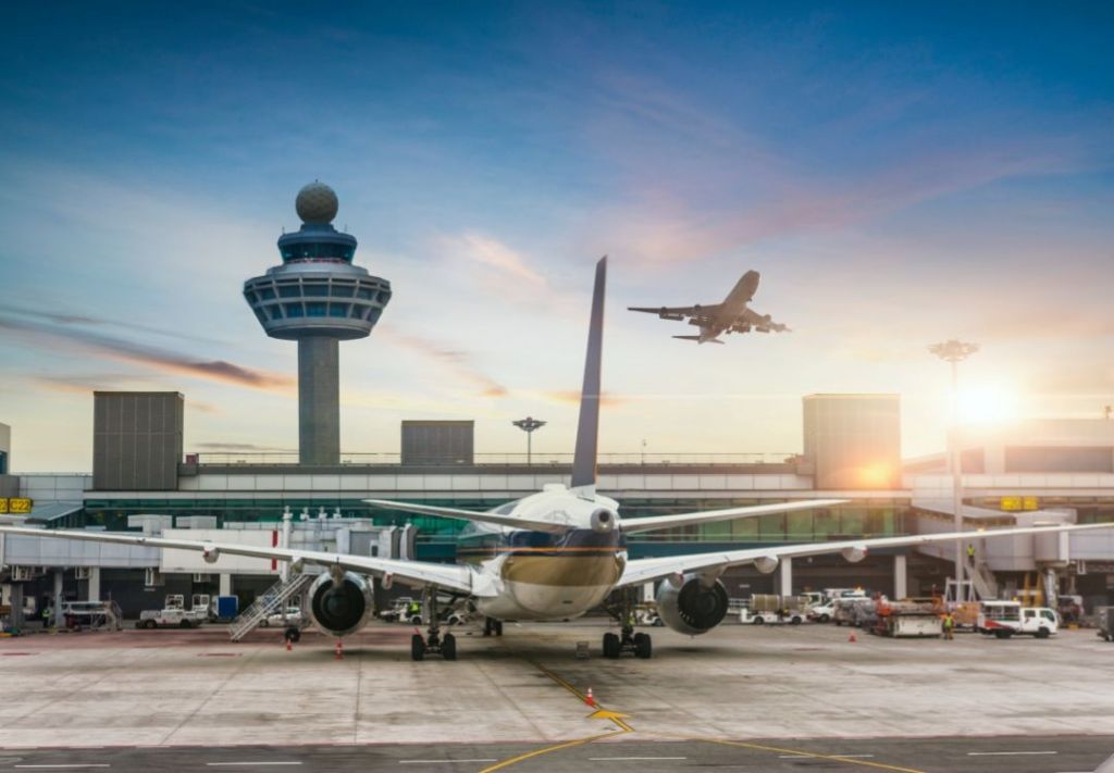 Sunset over a busy airport terminal with a commercial airliner taking off in the background and airport control tower visible.