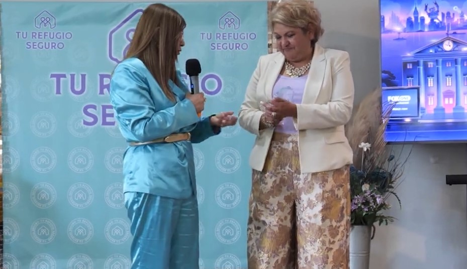 Two women on a stage, one interviewing the other in front of a "Tu Refugio Seguro" backdrop.