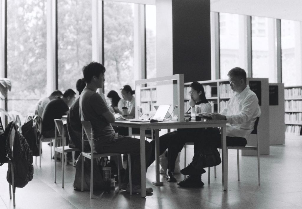 Black and white image of people working at desks in a library or study area, with bookshelves visible in the background.
