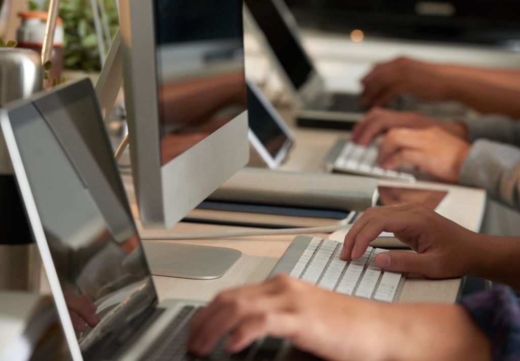 Hands typing on a keyboard in front of a computer monitor and other electronics on a desk.