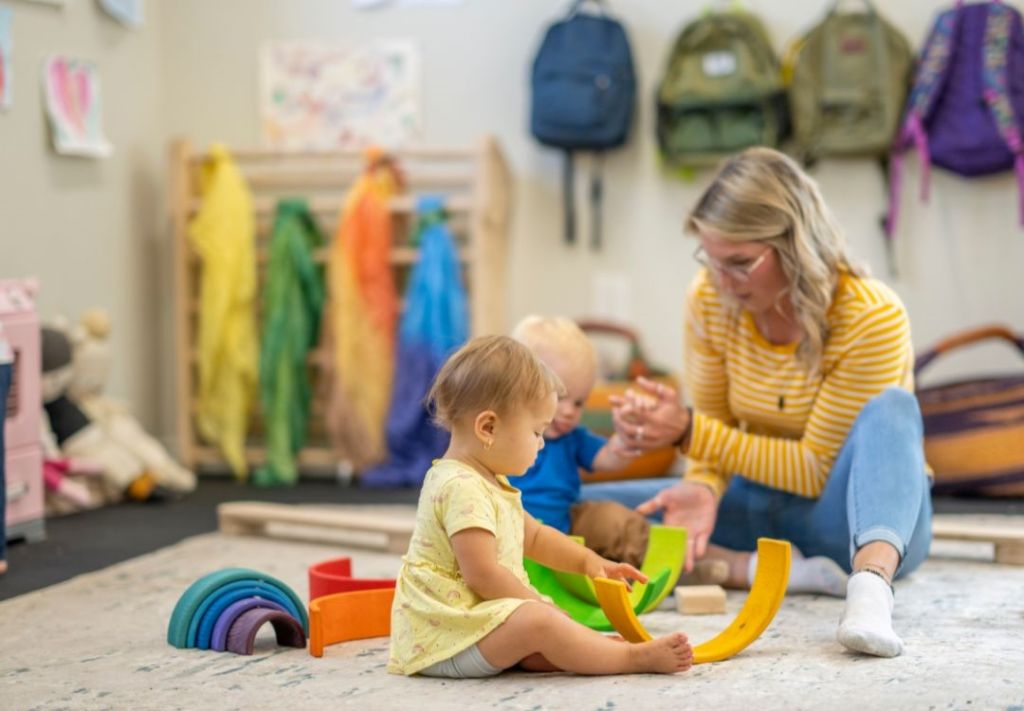 A woman in a yellow and white striped shirt sits on the floor playing with a young child in a daycare or preschool classroom setting, surrounded by colorful backpacks and toys.