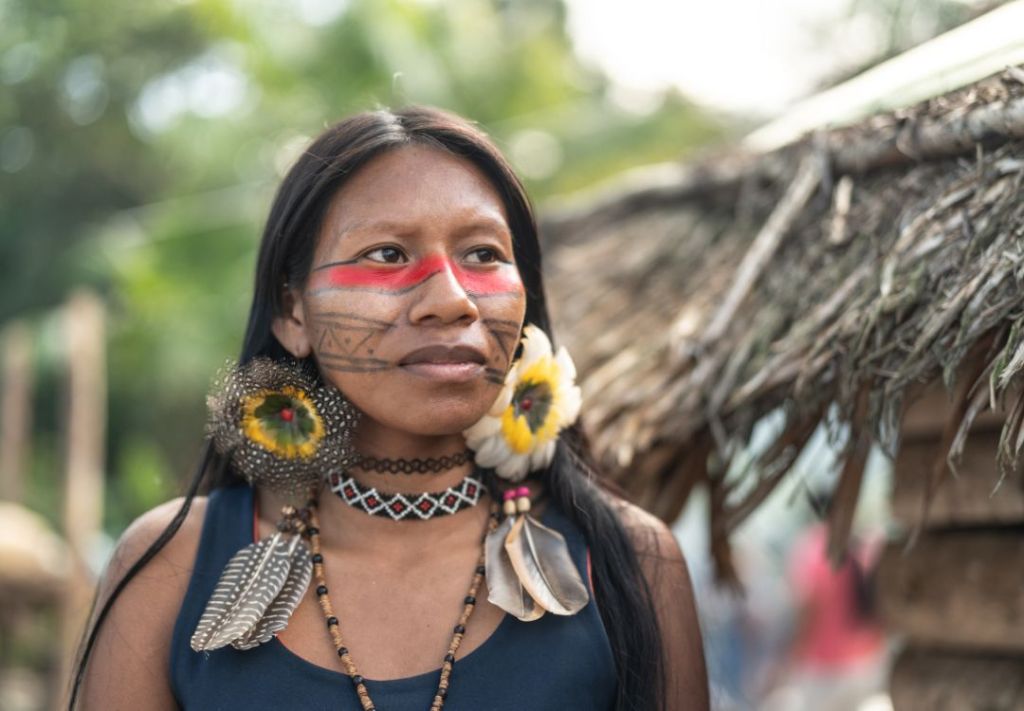 A woman with face paint and traditional jewelry stands in front of a thatched hut in a lush, green setting.