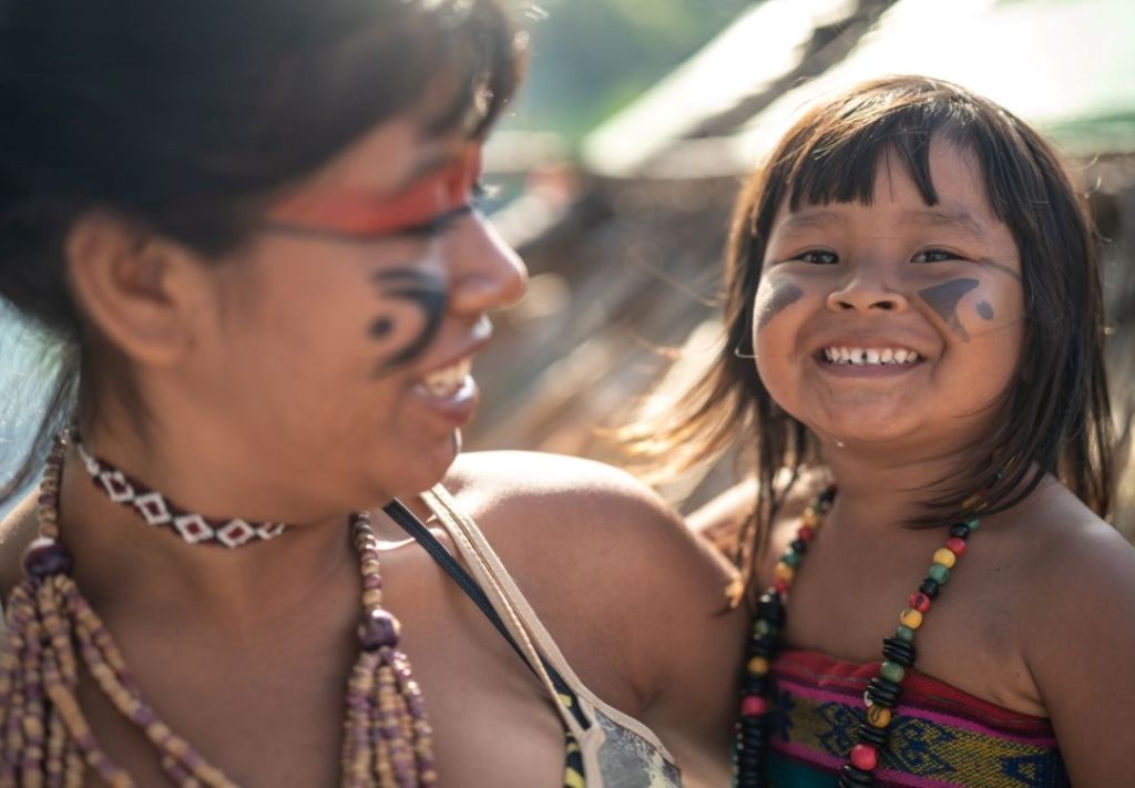 Two smiling Indigenous children wearing traditional beaded jewelry and face paint in a natural outdoor setting.