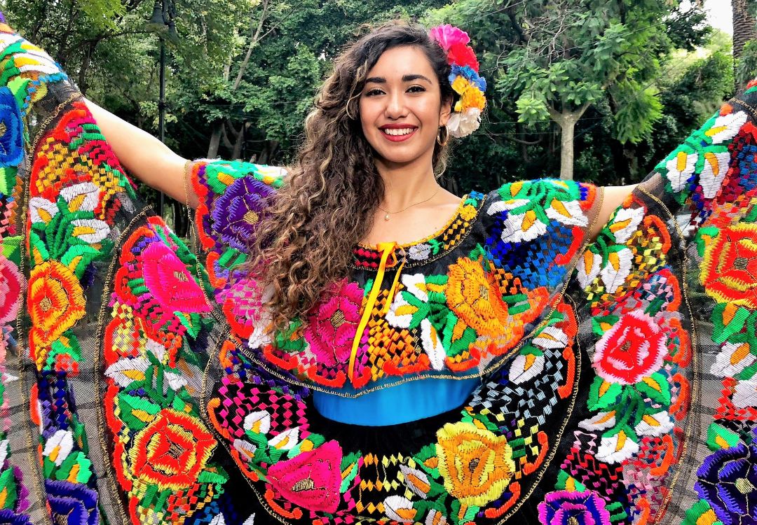 A smiling woman wearing a colorful, embroidered dress with floral patterns and holding her arms out in a joyful pose in a lush, green outdoor setting.