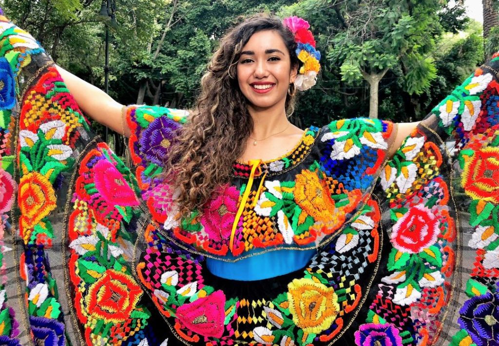 A smiling woman wearing a colorful, embroidered dress with floral patterns and holding her arms out in a joyful pose in a lush, green outdoor setting.