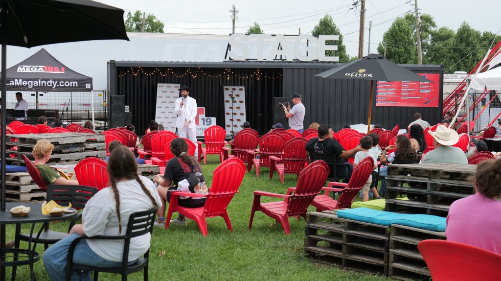 Outdoor food and entertainment event with red Adirondack chairs, food trucks, and a stage with performers.