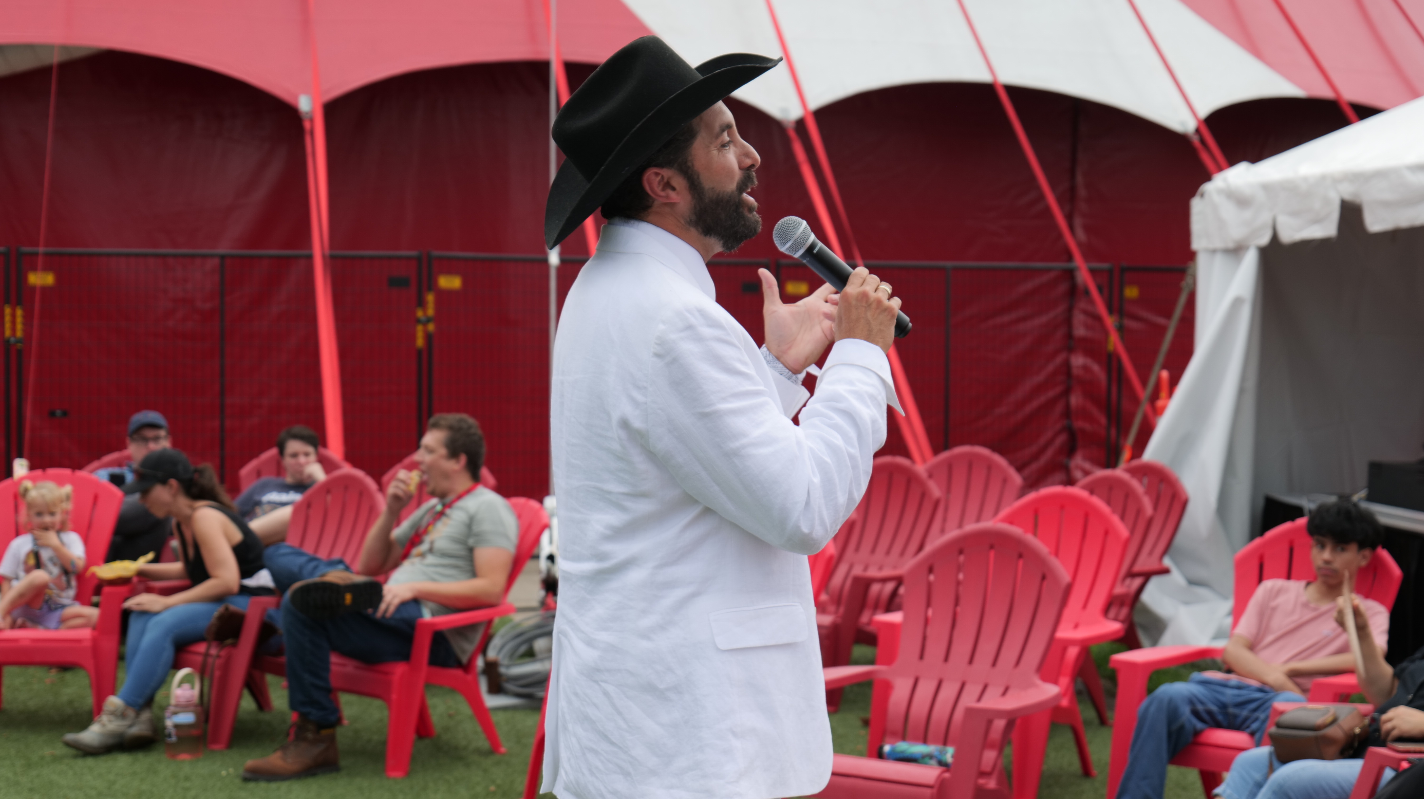 A man with a beard and cowboy hat speaking into a microphone at an outdoor event with red and white tents and chairs in the background.
