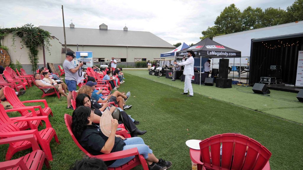 Outdoor event with people seated in red lawn chairs, tents and equipment on a grassy field, and a person speaking on a stage.