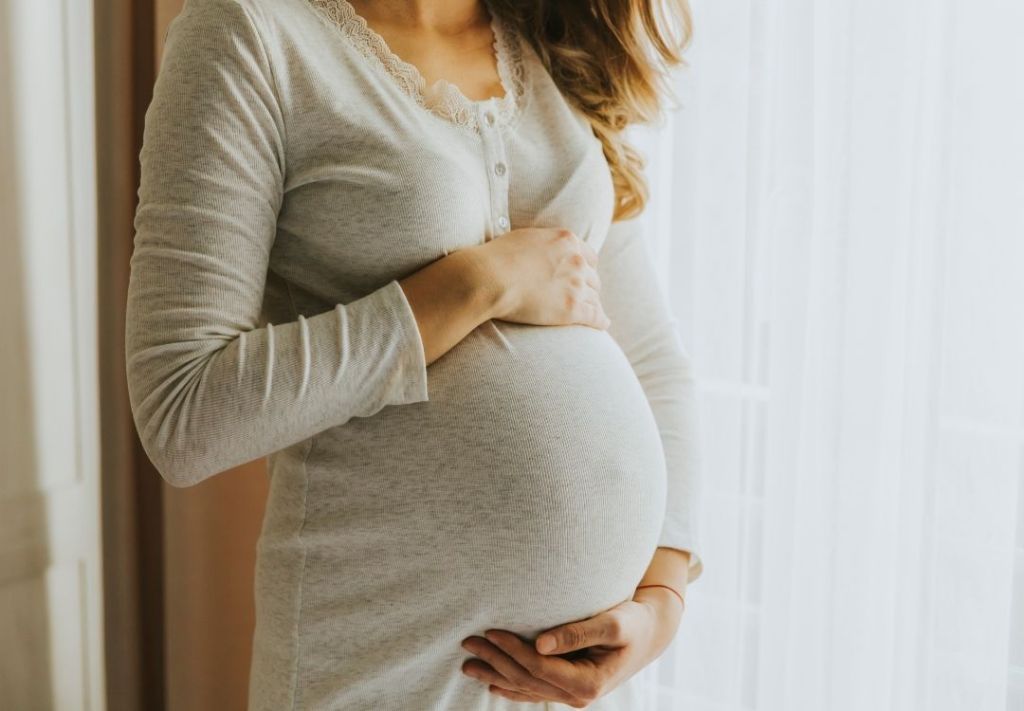 A pregnant woman wearing a gray sweater with lace trim, standing in a doorway and embracing her belly.