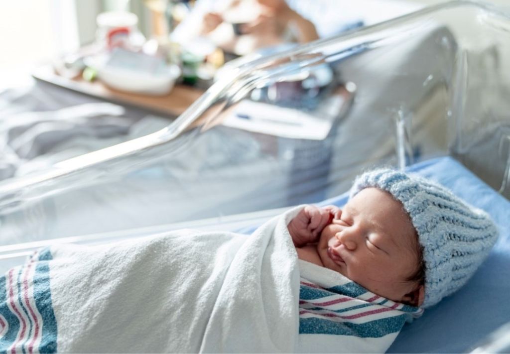 A sleeping newborn baby wrapped in a blue and white striped blanket, lying in a hospital bassinet.