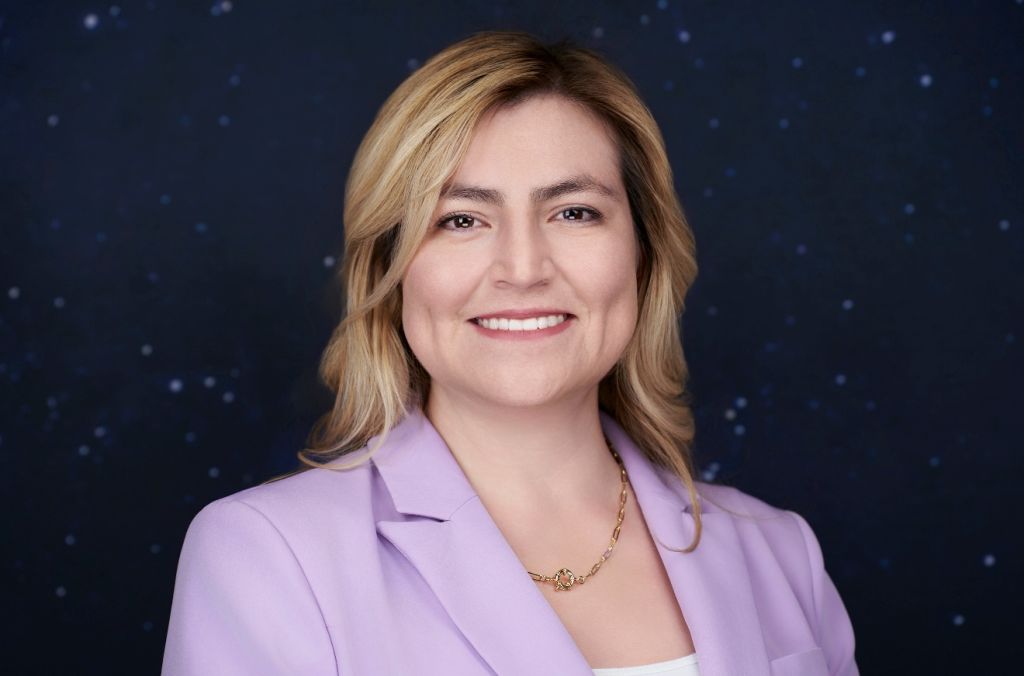 Smiling woman with blonde hair wearing a lavender blazer against a starry night sky background.