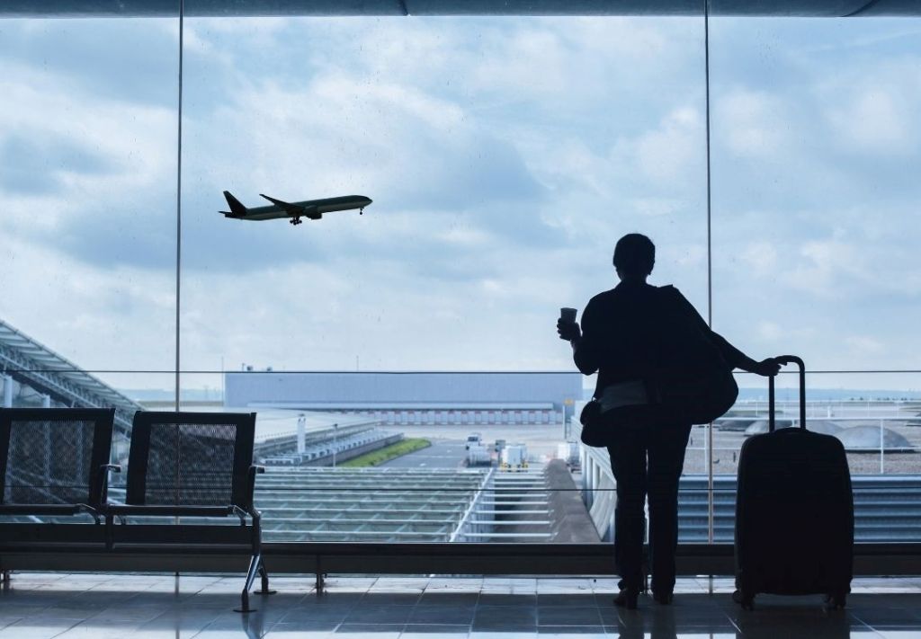 Silhouette of a person with luggage standing at an airport terminal, watching a plane take off in the distance through the window.