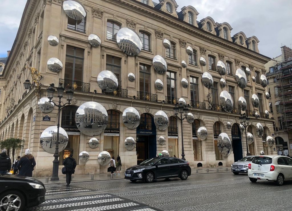 A grand, ornate building with numerous spherical light fixtures and the Louis Vuitton logo displayed prominently. Several vehicles are parked in front of the building.