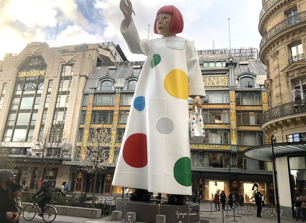 A large, colorful statue of a woman in a polka dot dress stands in a city square, surrounded by historic buildings and pedestrians.